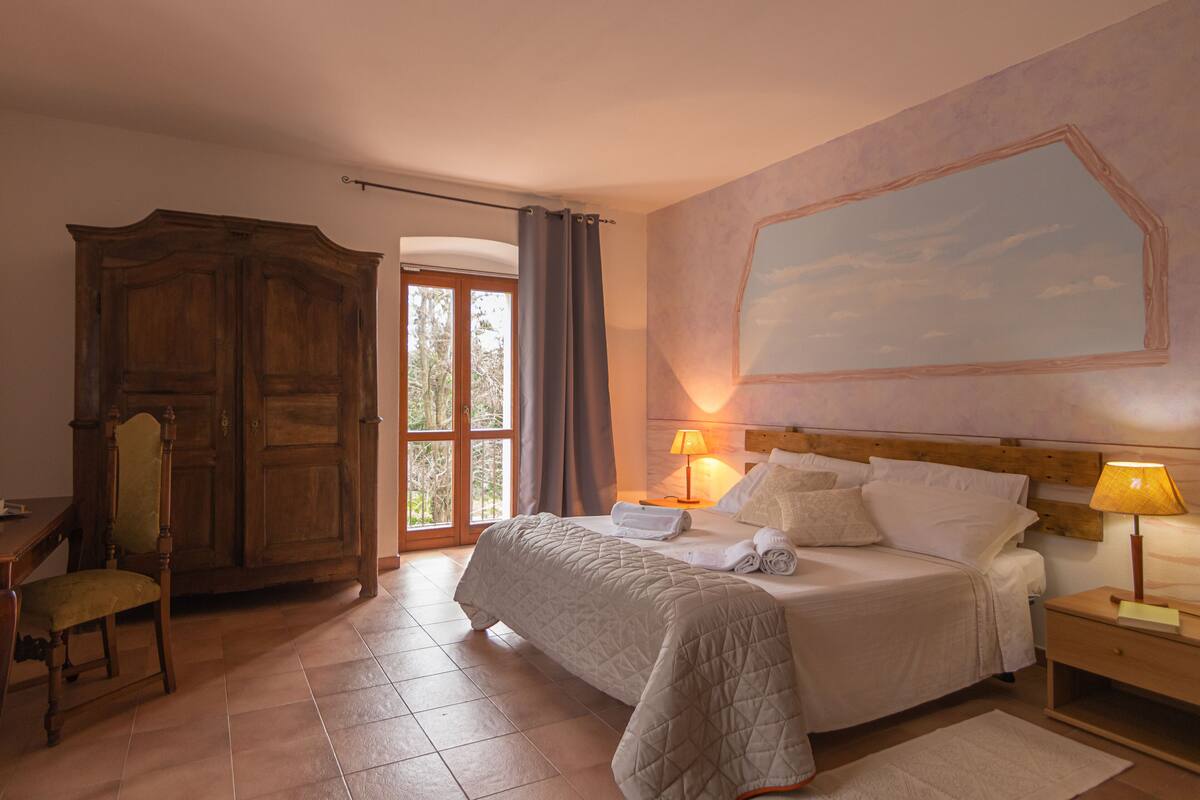 A bedroom with a large bed, a wooden wardrobe, a desk with a chair, and a window with curtains.