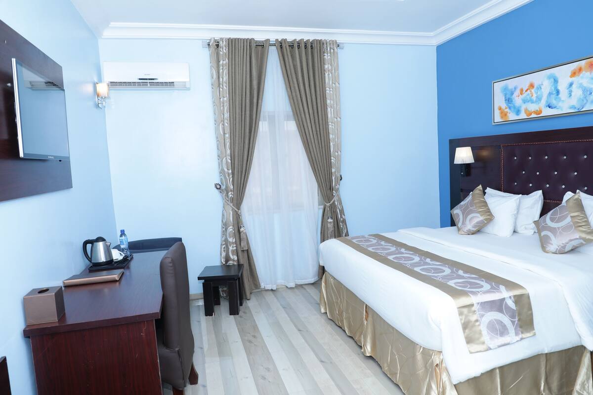 A hotel room with a bed, a desk, a chair, an air conditioning unit, and a painting on the wall.