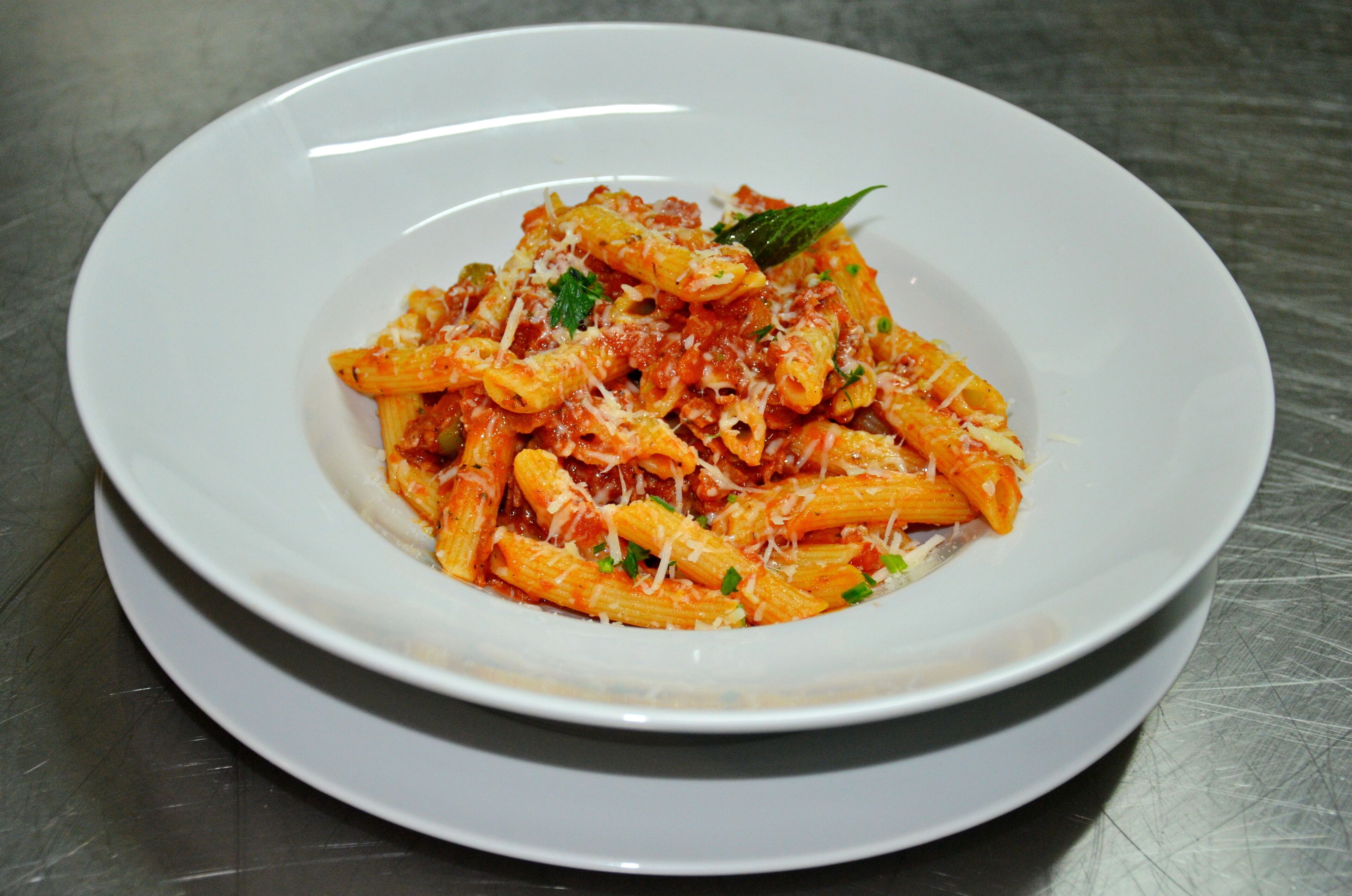 Une assiette blanche remplie de pâtes penne nappées de sauce tomate et de fromage, posée sur une table en acier inoxydable.
