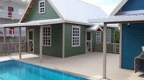 Uma área junto à piscina com um edifício verde, um deque coberto e um edifício azul ao fundo.