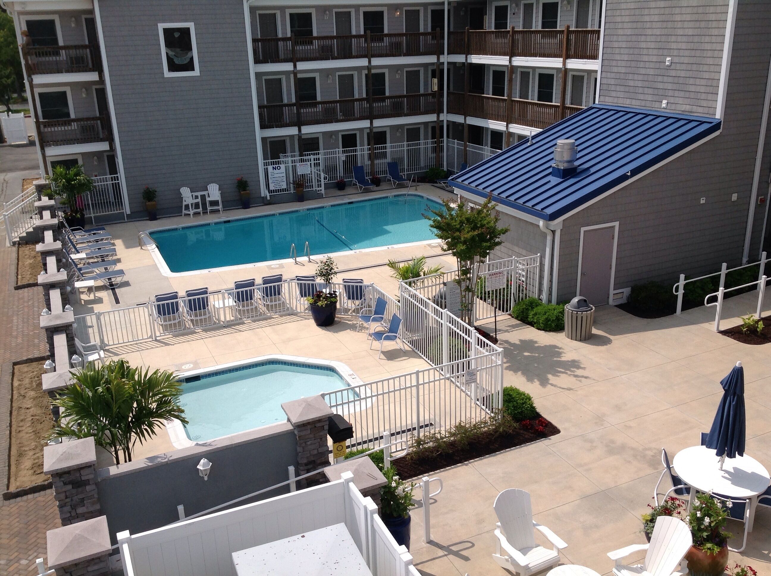 A rooftop pool area with multiple lounge chairs and a smaller pool, surrounded by a fence and a building with balconies.