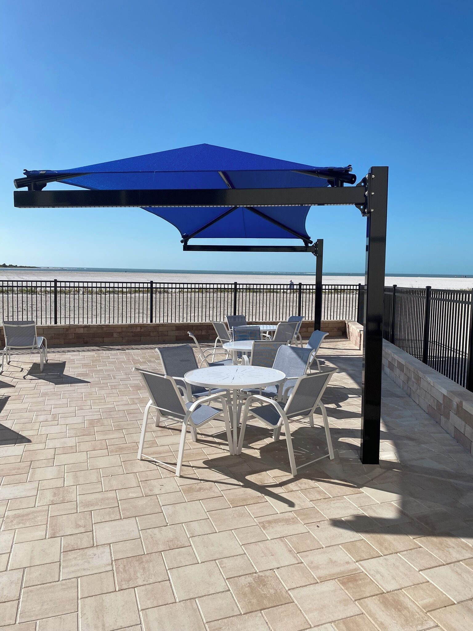 An outdoor patio with a blue canopy, white round tables, and metal chairs.