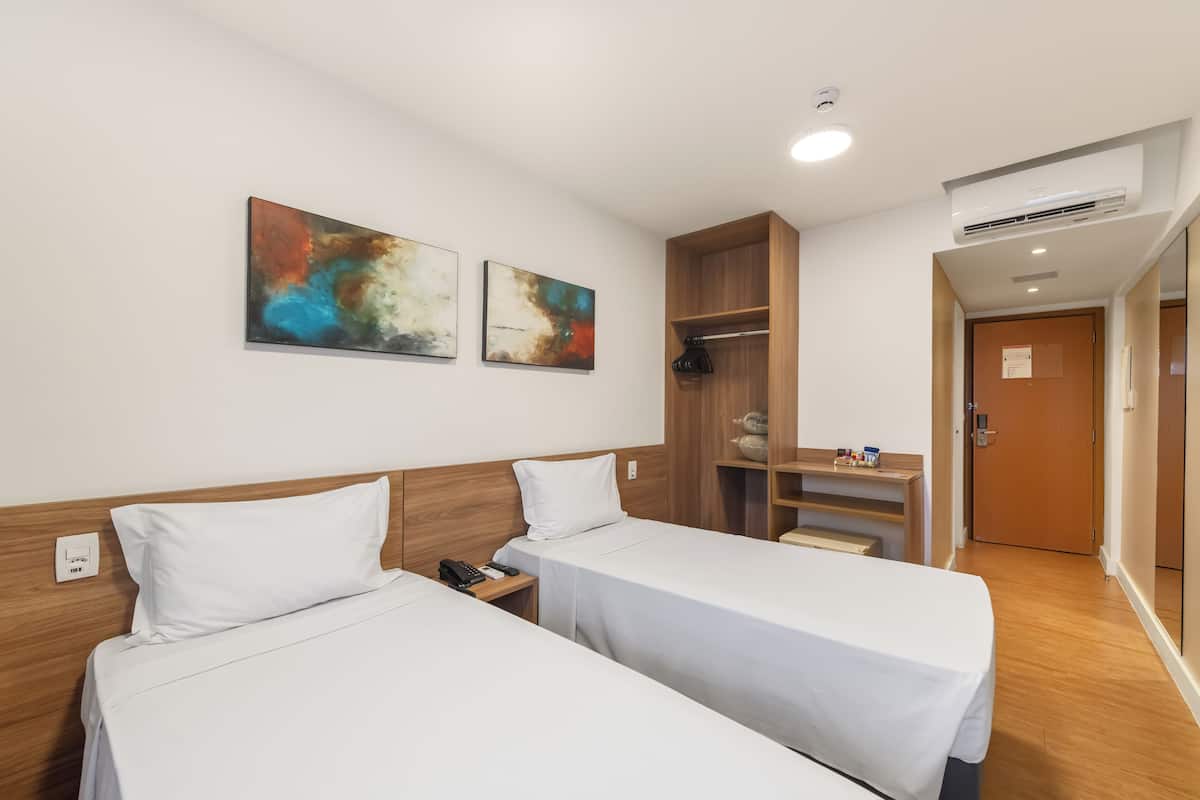 A hotel room with two single beds, a wooden headboard, a desk with a chair, a wardrobe, and abstract wall art.