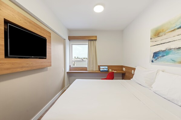 A hotel room with a large bed, a mounted TV, a window with curtains, a desk with a laptop, and a painting on the wall.