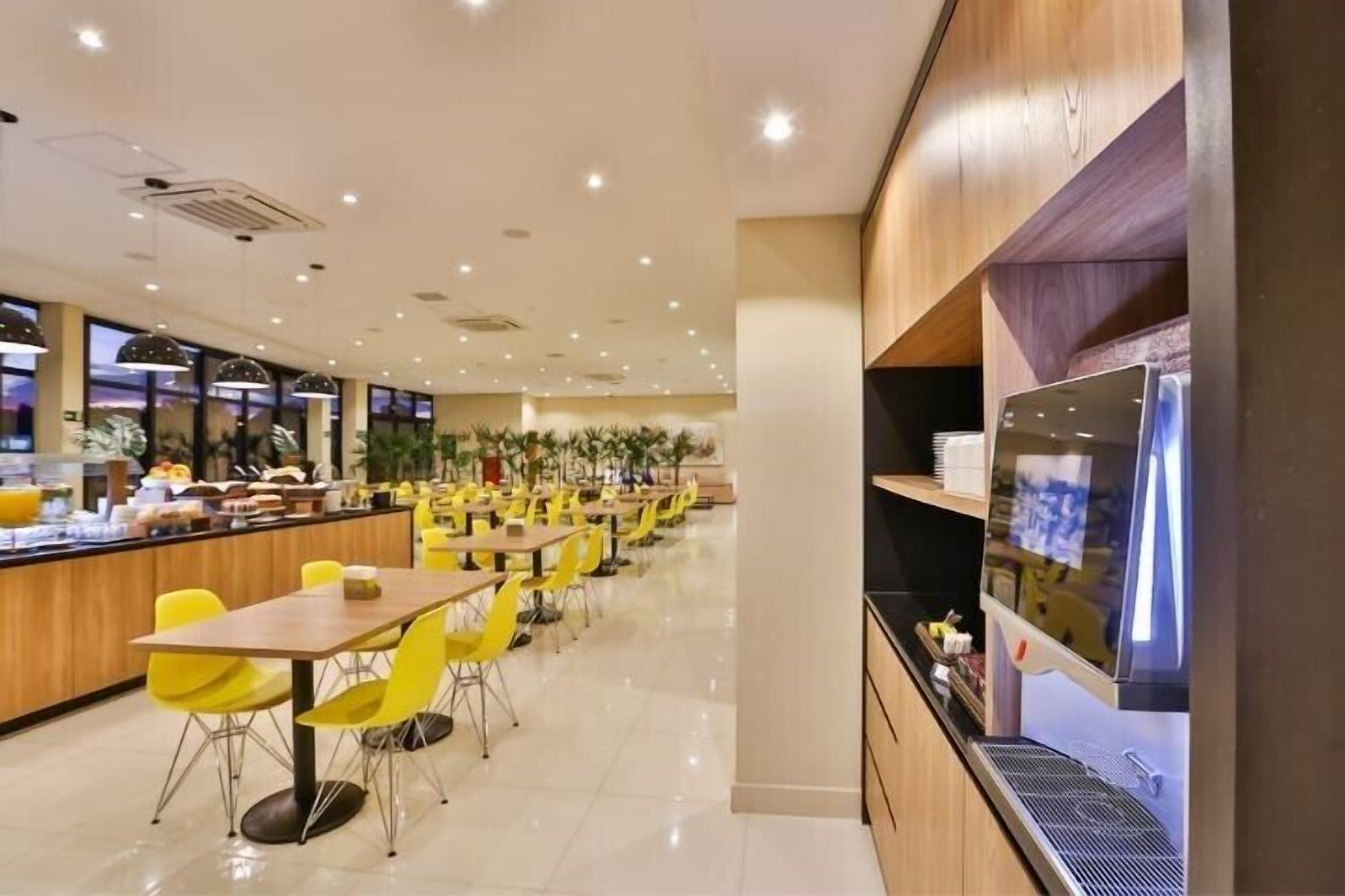 A modern dining area with yellow chairs and tables, a counter with food items, and a large screen on the wall.