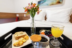 Ett brickbord med croissanter, en våffla, kaffe och en skål med frukt står på sängen i ett hotellrum.