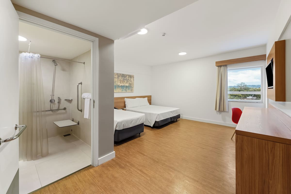 A hotel room with a shower, two beds, a small sink, a wooden desk, a red chair, and a window with a view.