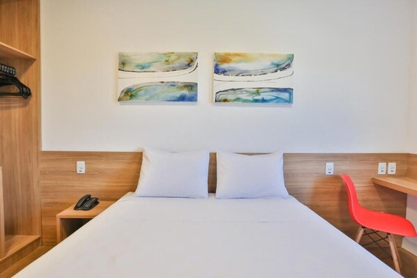 Habitación de hotel con una cama, dos almohadas, una silla roja, una mesita de noche de madera con un teléfono y tres cuadros abstractos en la pared.