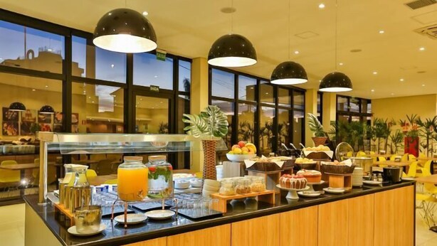 A buffet counter with various food items and a fruit bowl, surrounded by large windows and indoor plants. - Blue Tree Jaguariuna (Jaguariuna)