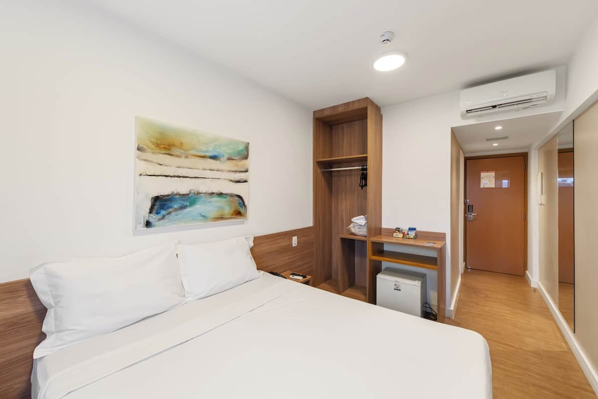 A hotel room with a bed, a wooden wardrobe, a small desk with a chair, and an air conditioning unit.