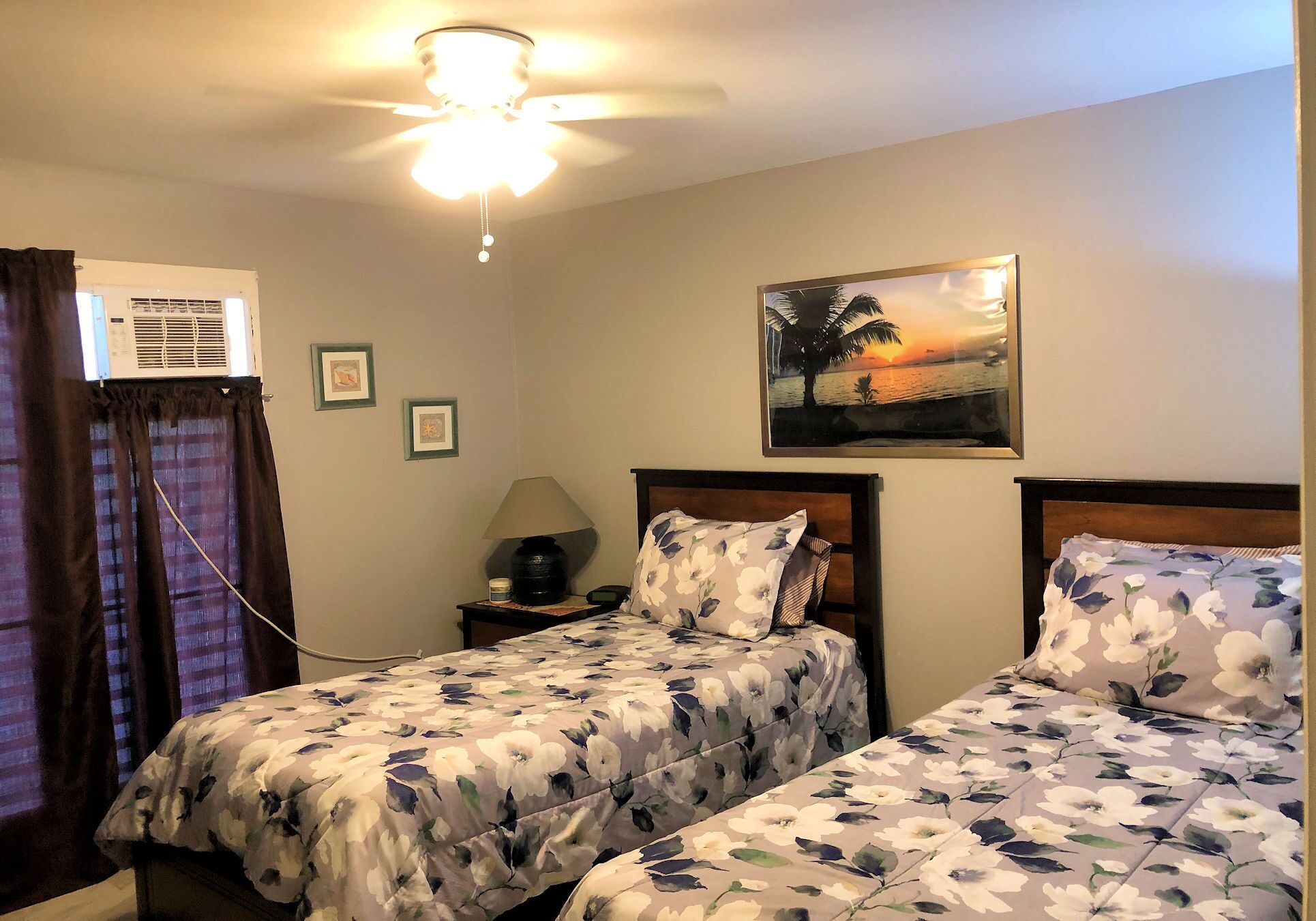 Une chambre d’hôtel avec deux lits, un ventilateur de plafond, un climatiseur, un tableau représentant un coucher de soleil et une table de chevet avec une lampe.