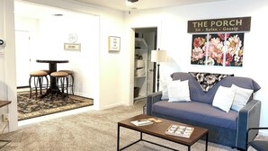 A living room with a sofa, coffee table, and a ceiling fan. There is a sign on the wall that reads "THE PORCH Sit Relax Gossip".