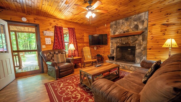 A cozy cabin interior with a stone fireplace, wooden walls, a leather couch, and a television mounted on the wall.