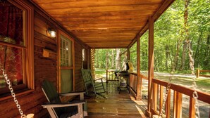 A wooden porch with a view of a forest. There are two rocking chairs and a table with chairs.