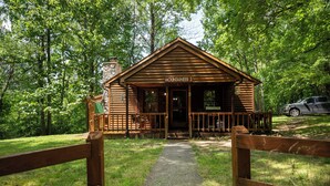 A wooden cabin named "Mountaineer 3" with a porch and a stone chimney, surrounded by a wooded area.