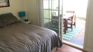 Un dormitorio con cama, mesita de noche, lámpara y puerta corrediza de vidrio que da a un patio exterior con mesa y sillas.