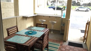 Un balcón con una mesa y sillas de madera, un taburete pequeño y vistas a la calle con coches y palmeras.