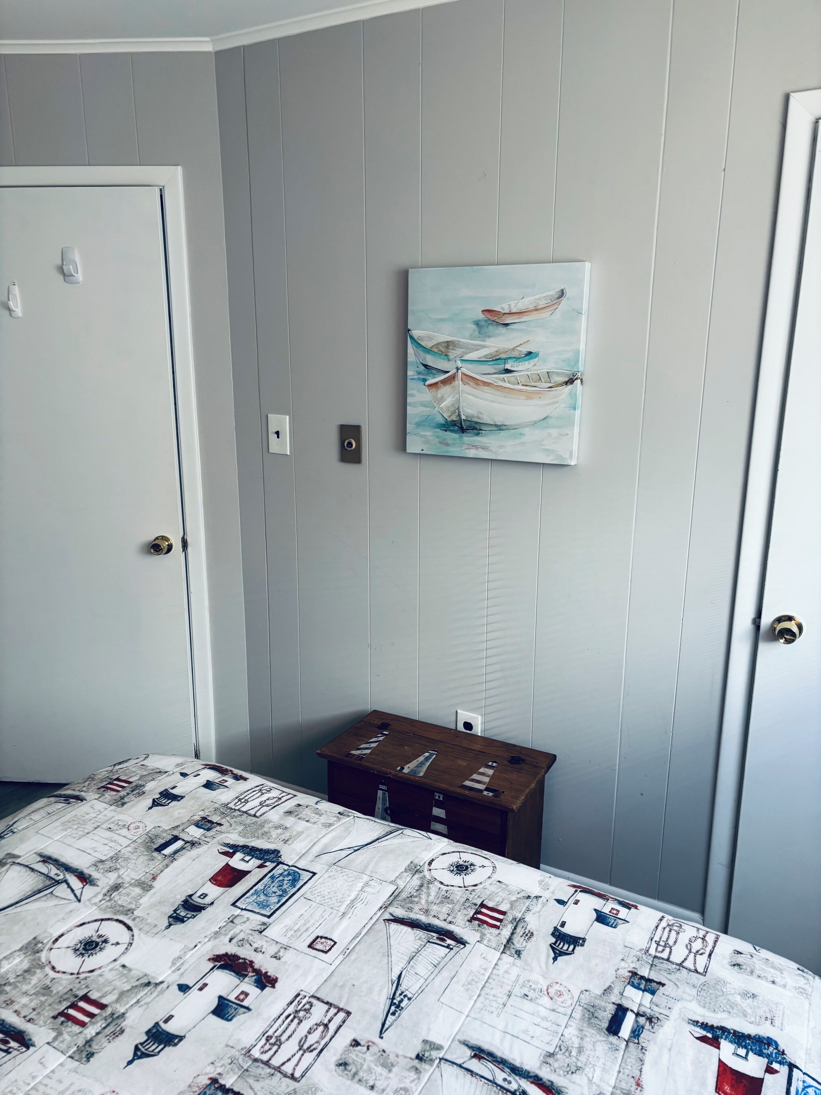 Quarto com cama, pintura de barcos, pequena mesa de madeira e porta branca.