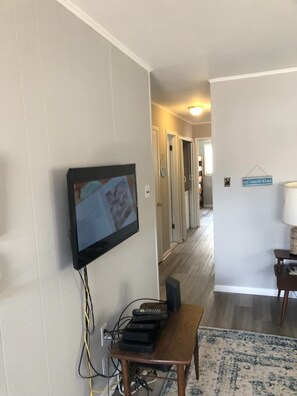 A hallway with a mounted television displaying a beach scene, a wooden table with electronic devices, and a rug on the floor.
