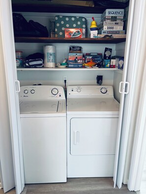 A laundry room with two front-loading washing machines, shelves with cleaning supplies, and a sequence game box. - Perfect Vacation Summer Spot in LBI NJ - first floor (Surf City)