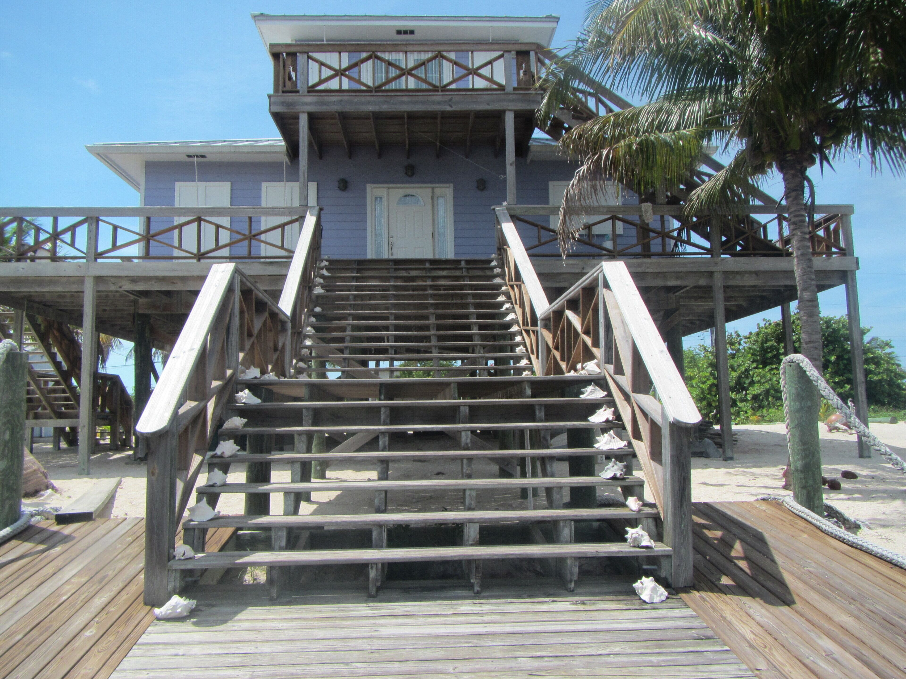 Una escalera de madera que conduce a un edificio con balcón, rodeado de palmeras y una playa de arena.