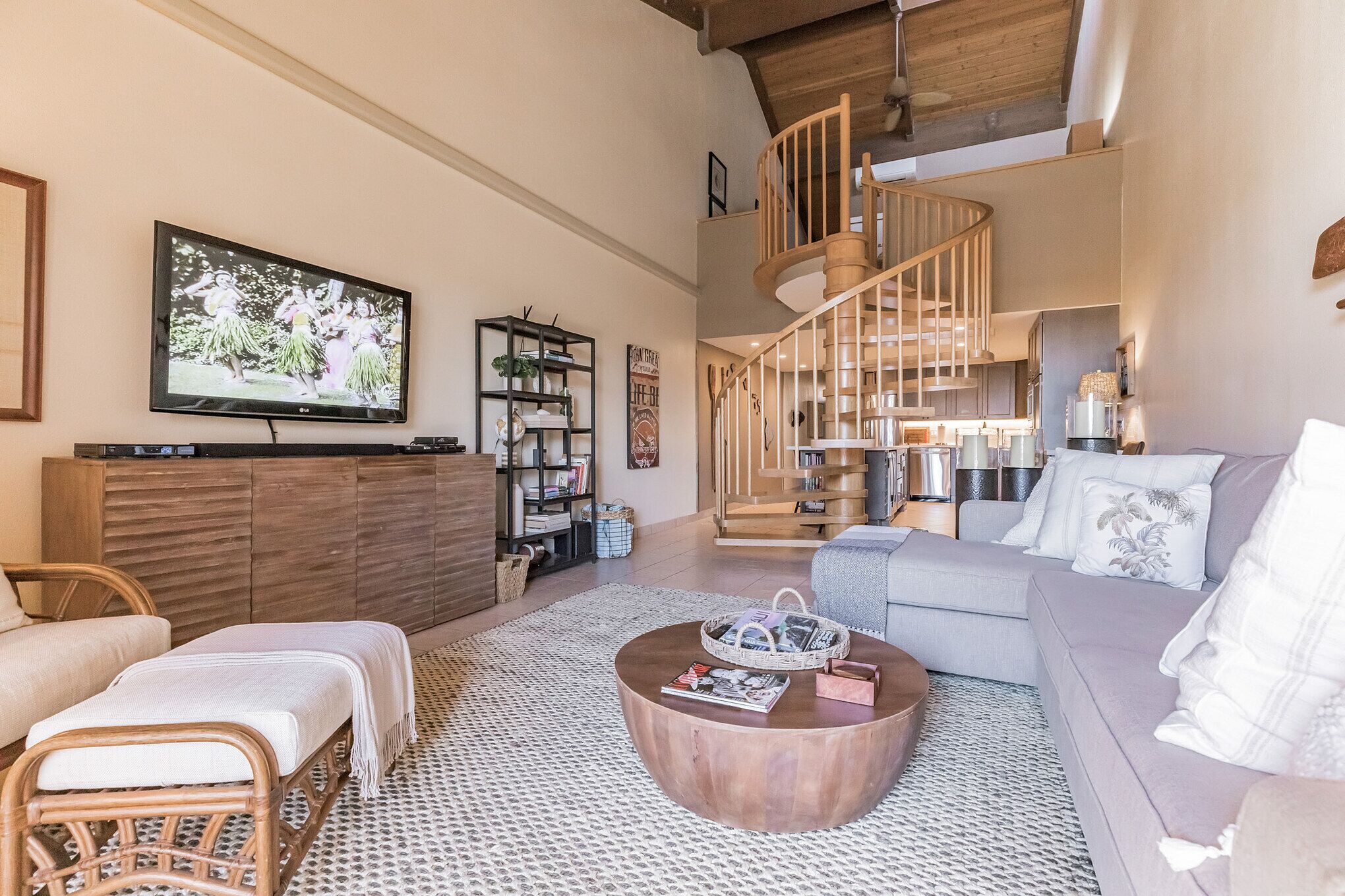 Amplia sala de estar con una mesa de centro de madera, un sofá gris, un sillón de mimbre, un televisor montado en la pared y una escalera de caracol que conduce a un nivel superior.