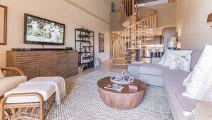 Un vaste espace de vie comprenant une table basse en bois, un canapé gris, un fauteuil en osier, un téléviseur fixé au mur et un escalier en colimaçon menant à un niveau supérieur.