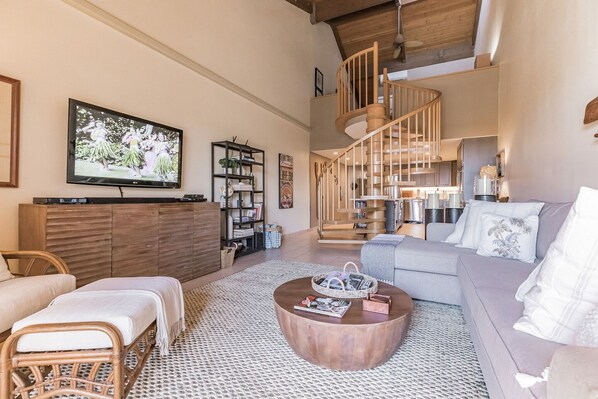 A spacious living area with a wooden coffee table, a grey sofa, a wicker chair, a TV mounted on the wall, and a spiral staircase leading to an upper level.