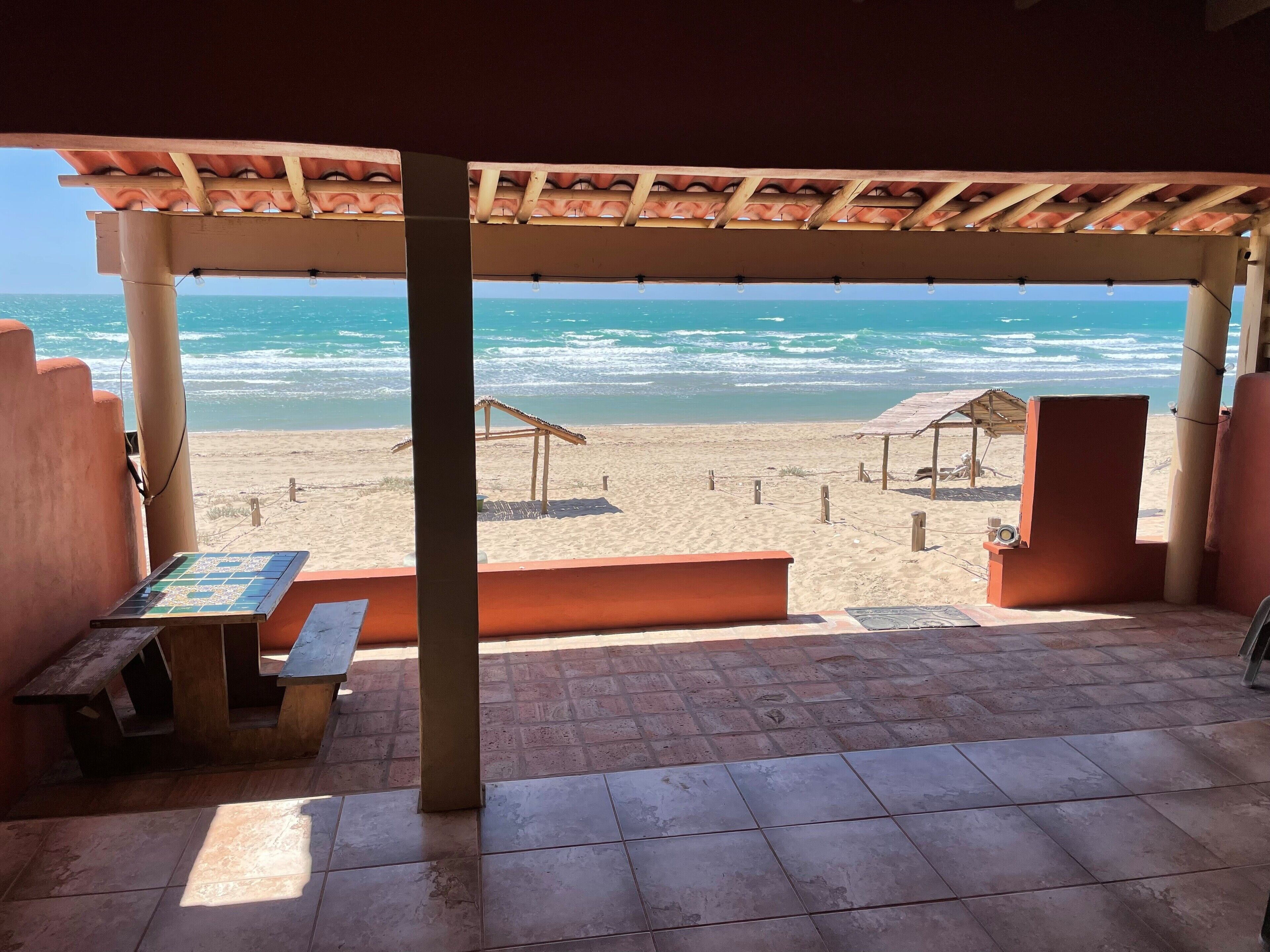 A beachside patio with a view of the ocean, featuring a tiled floor, wooden benches, and a pergola with a tiled roof.