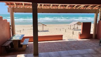 A beachside patio with a view of the ocean, featuring a tiled floor, wooden benches, and a pergola with a tiled roof.