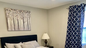 Ein Schlafzimmer mit einem Bett, einem Nachttisch, einer Lampe, einem Gemälde und einem gemusterten Vorhang.