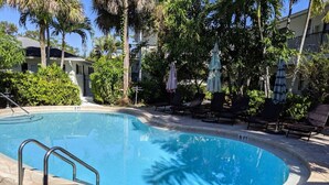 Une piscine dont la profondeur est indiquée à 6 pieds, entourée de chaises longues et de parasols, avec des palmiers et un bâtiment en arrière-plan.
