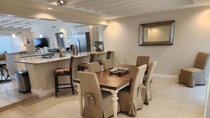 Un coin repas avec une table en bois et six chaises, une cuisine à l’arrière-plan et un grand miroir au mur.