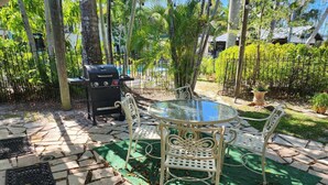 Une terrasse extérieure avec une table en verre entourée de chaises en métal blanc, un barbecue et une plante en pot.