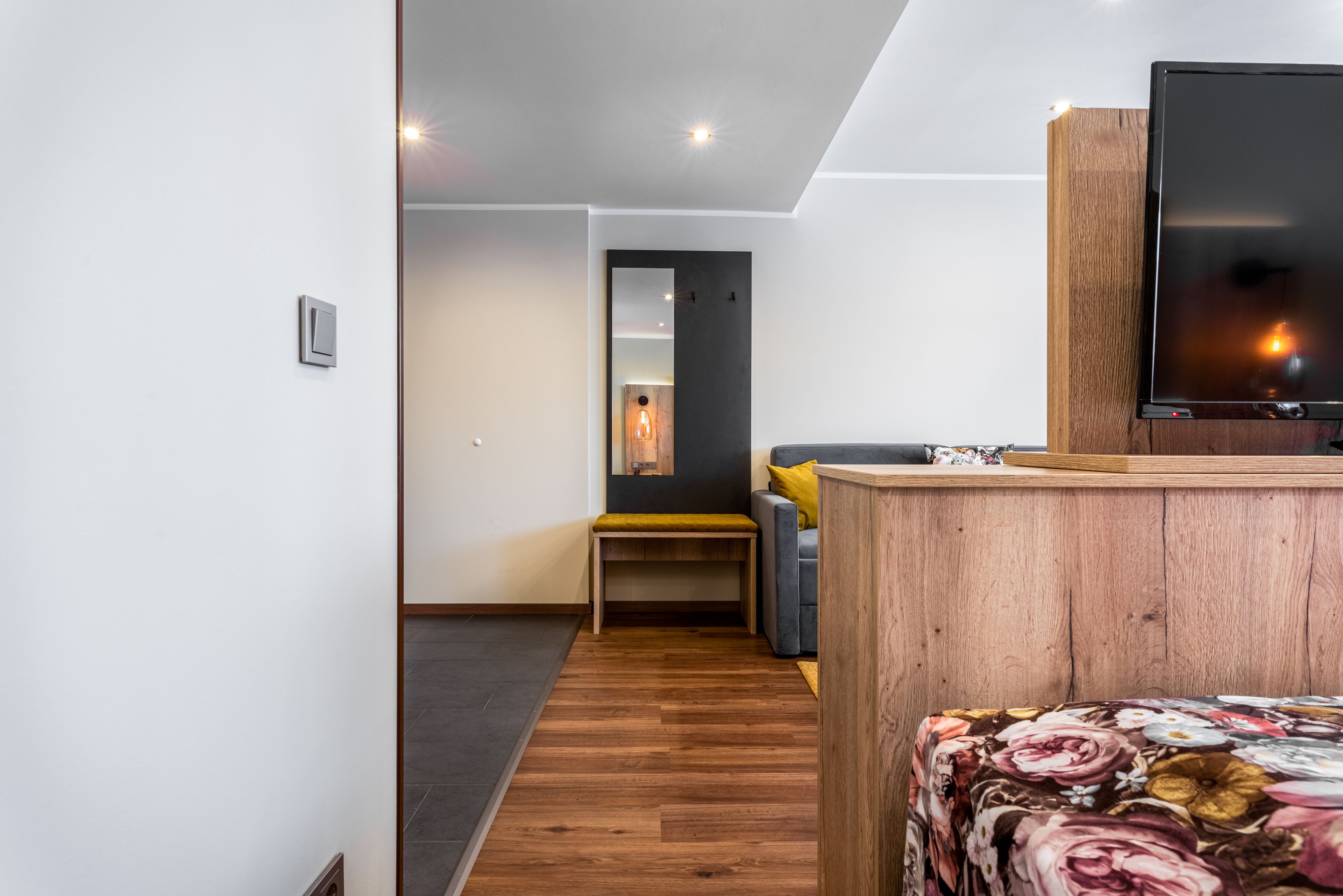 Moderni hotellihuone, jossa on puinen työpöytä, litteänäyttöinen televisio, keltainen koristetyynyillä varustettu sohva ja penkki.