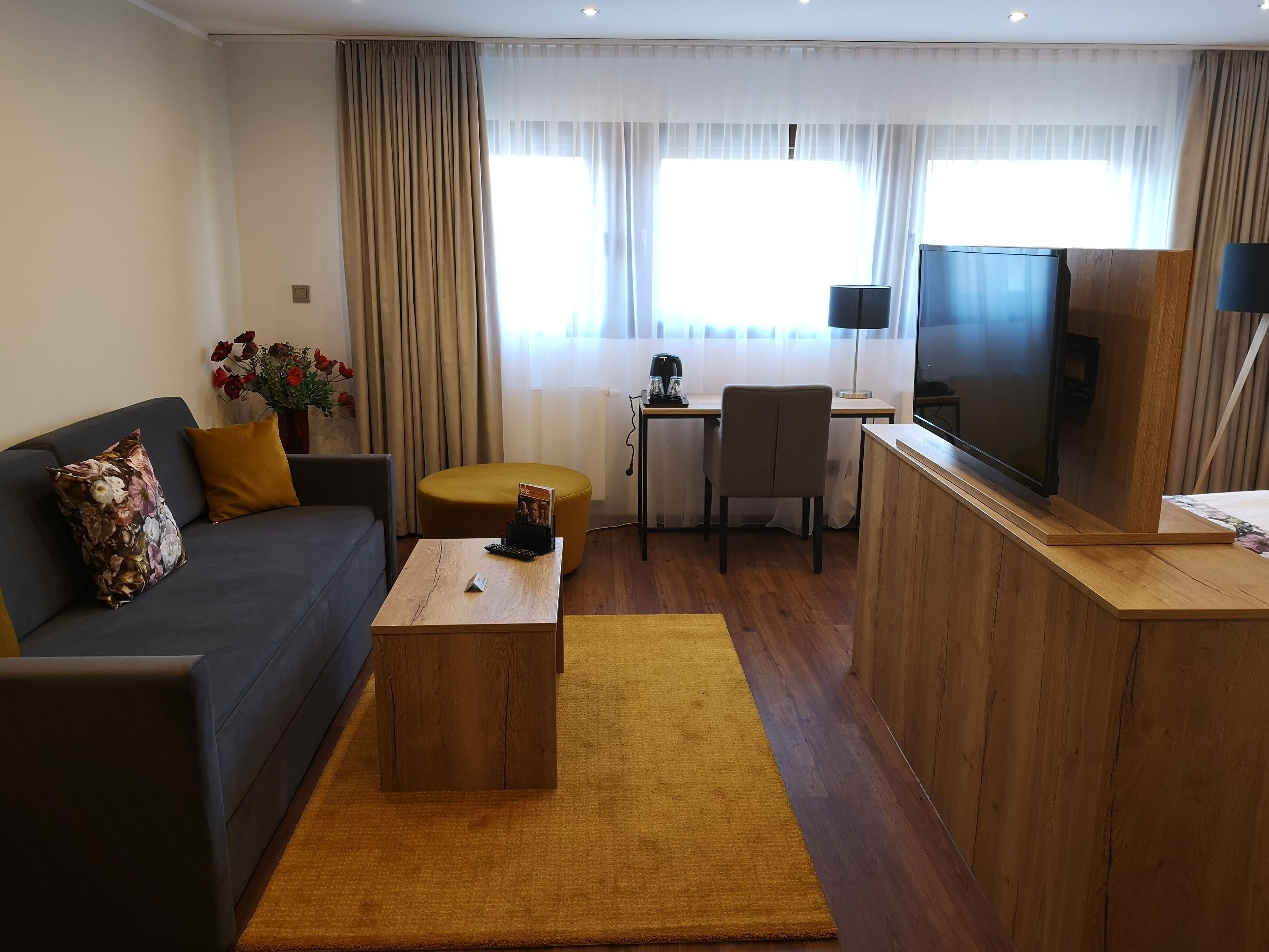 A modern living room with a sofa, coffee table, and a TV. There is a small table with a remote control and a magazine. The room has wooden flooring and beige curtains.