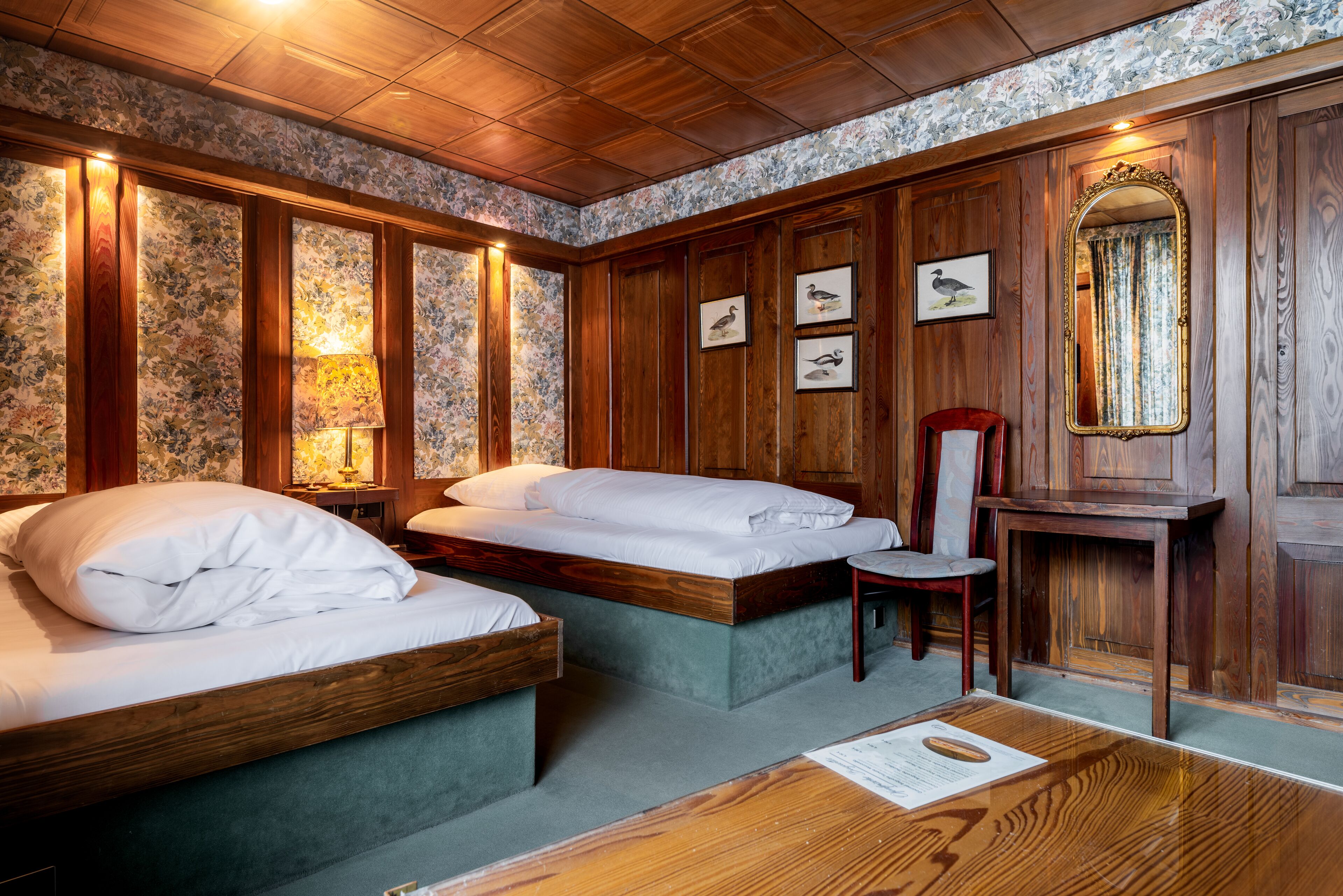 A room with two beds, a wooden desk, a chair, and framed pictures on the wall.