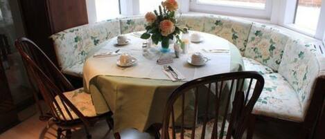Mesa redonda con un sofá floral, rodeada de sillas de madera, preparada para una comida con tazas de té y un florero.