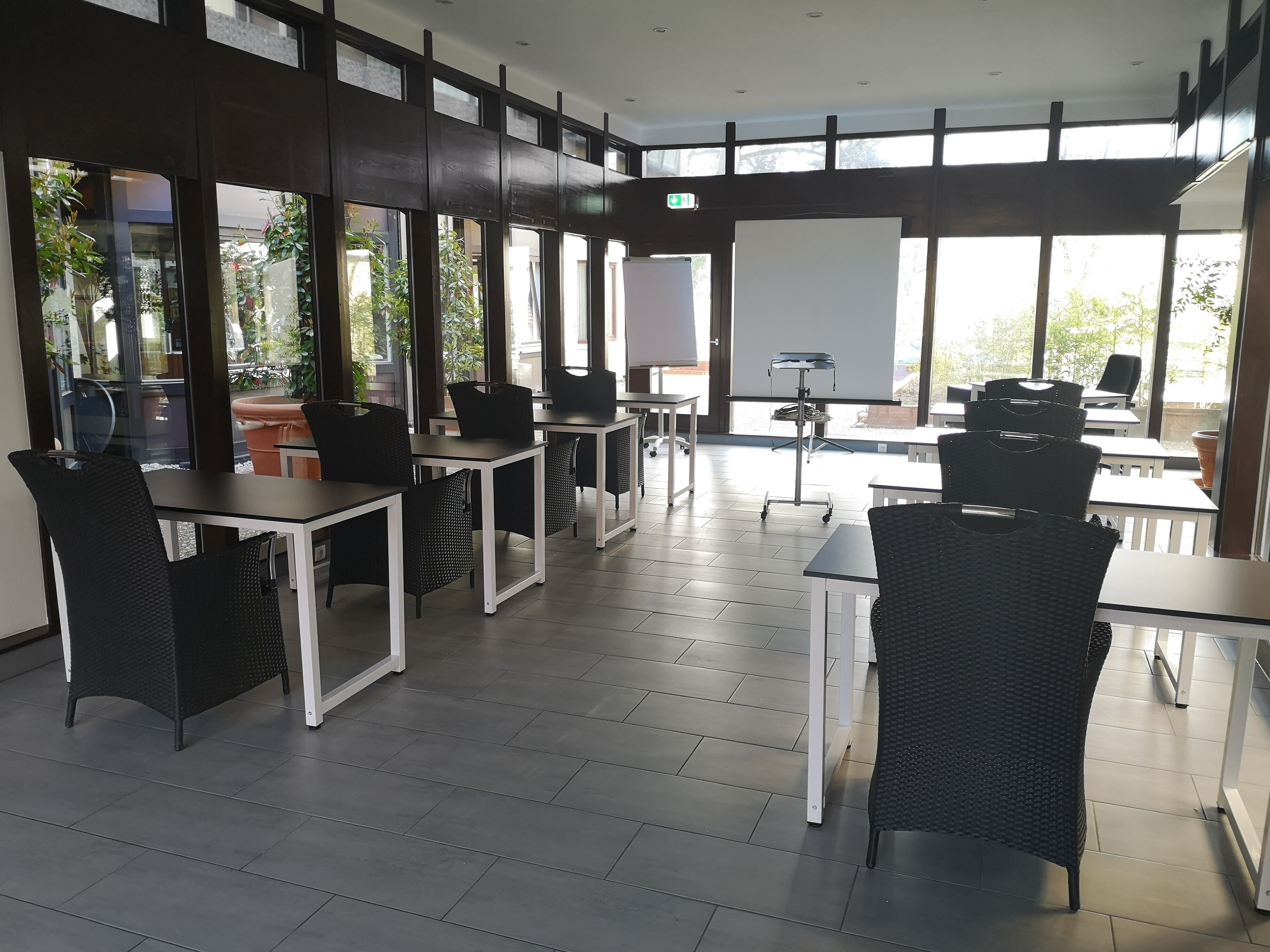 A modern indoor dining area with black wicker chairs and white tables, large windows offering views of greenery outside.