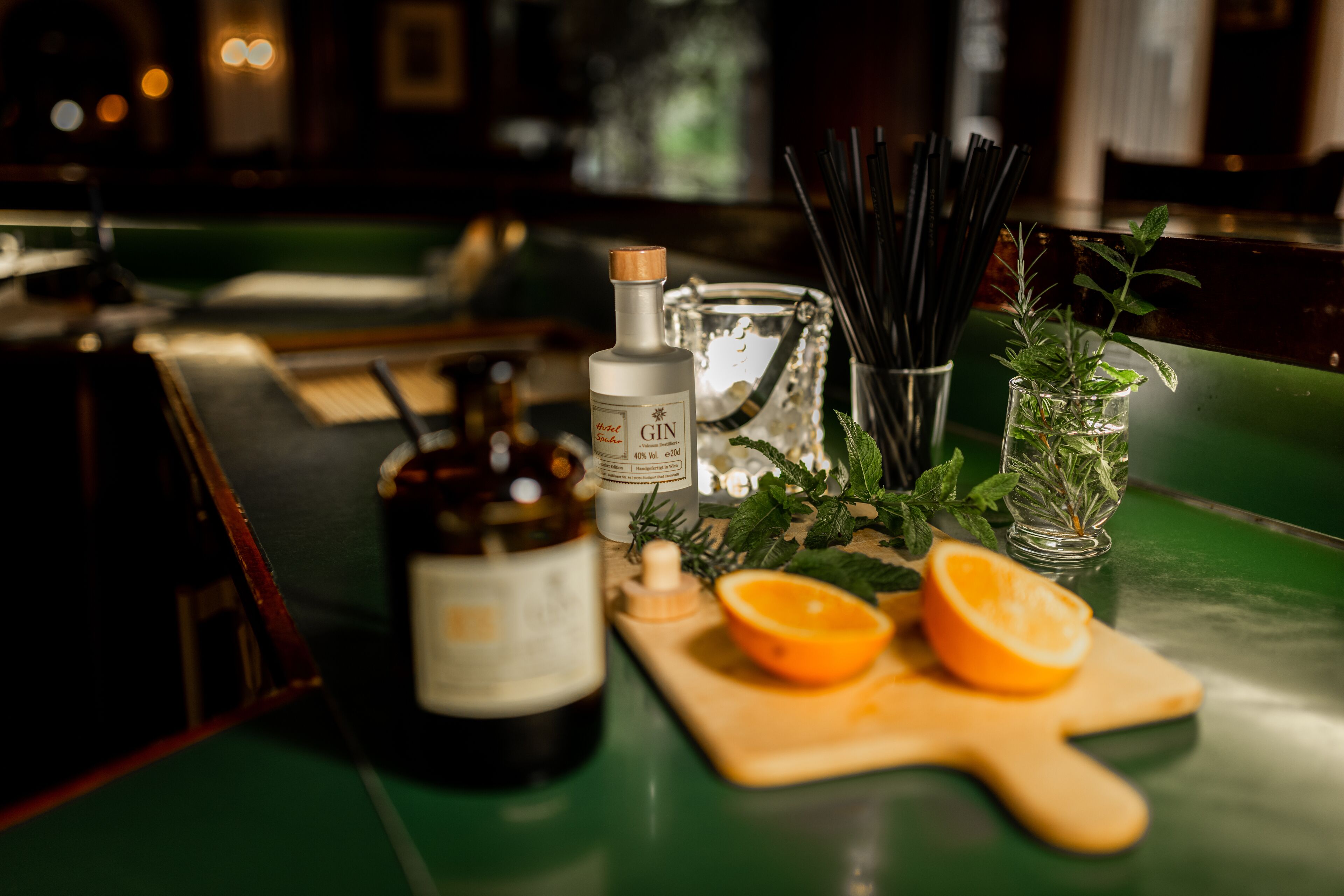 Eine Bar-Szene mit einer Gin-Flasche, einem Glas Wasser, einem Holzbruchbrett mit einer Orangenscheibe und einem Glas mit Kräutern.