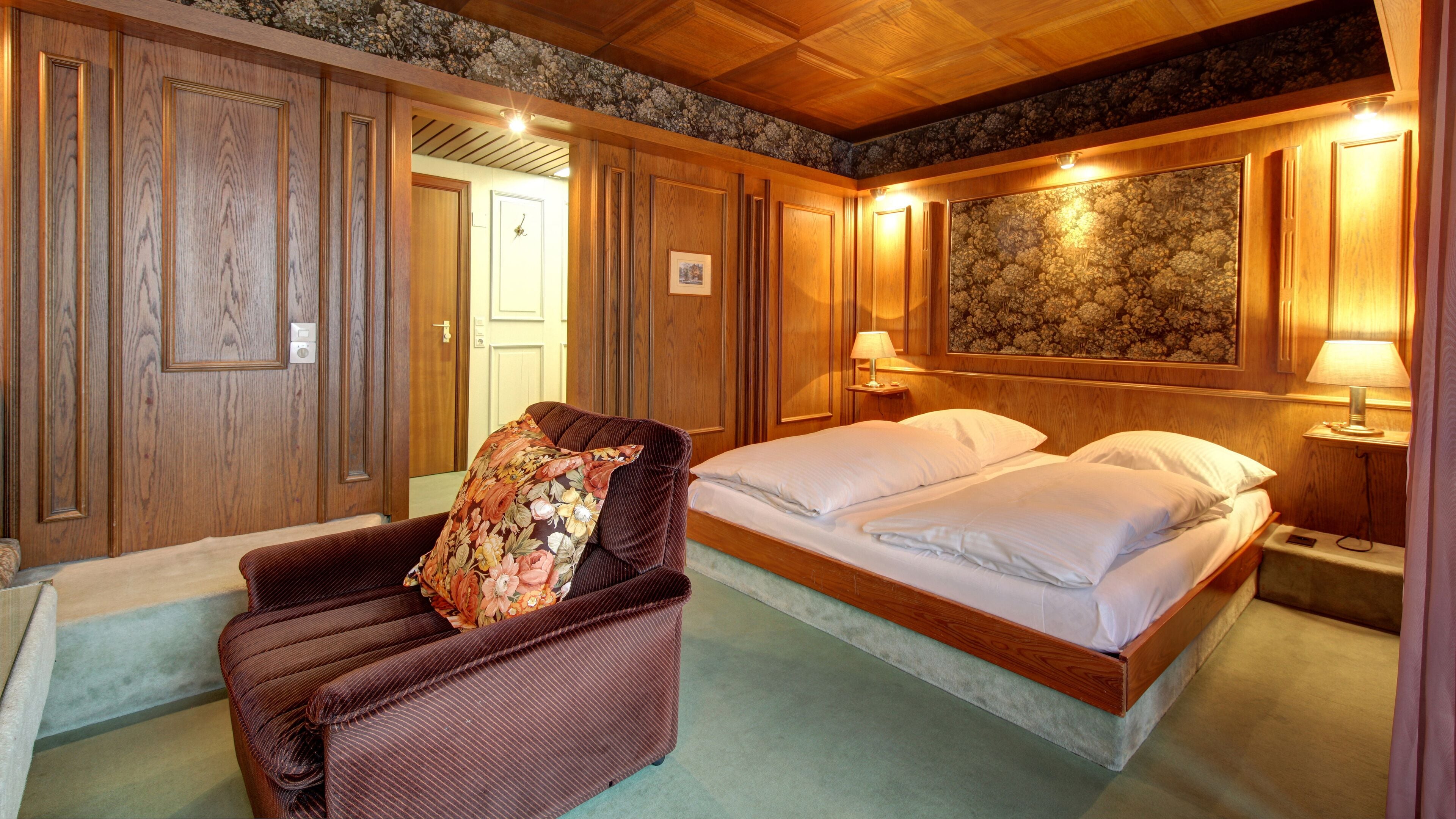 Una habitación de hotel con una cama de madera, una silla con cojín estampado y una gran obra de arte floral en la pared.