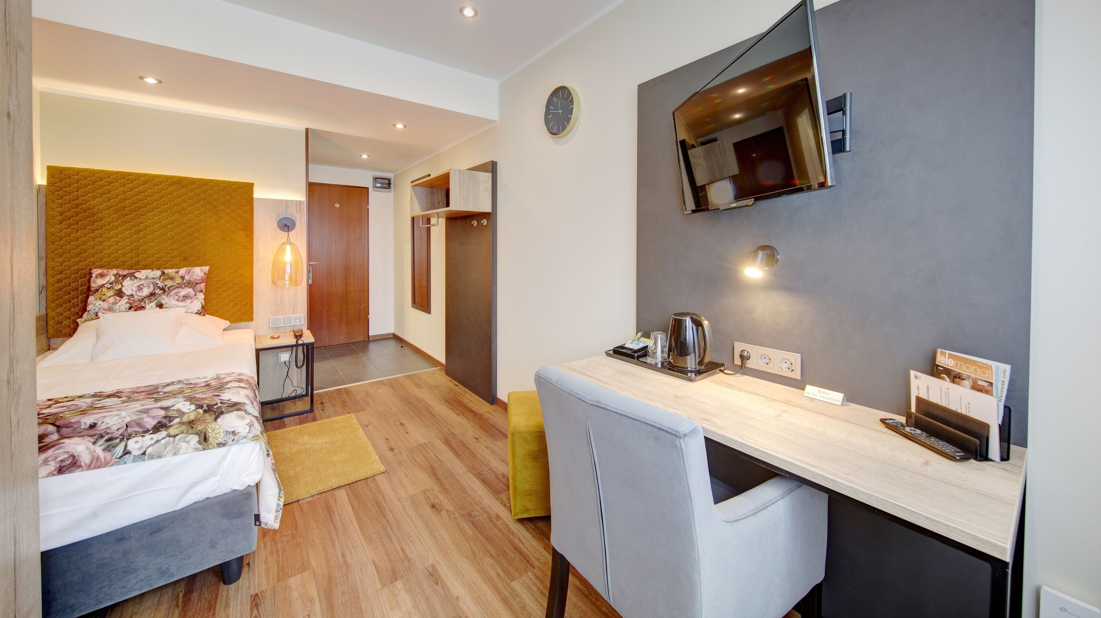 Une chambre d’hôtel moderne équipée d’un lit, d’un bureau, d’une chaise, d’un miroir et d’une horloge murale.