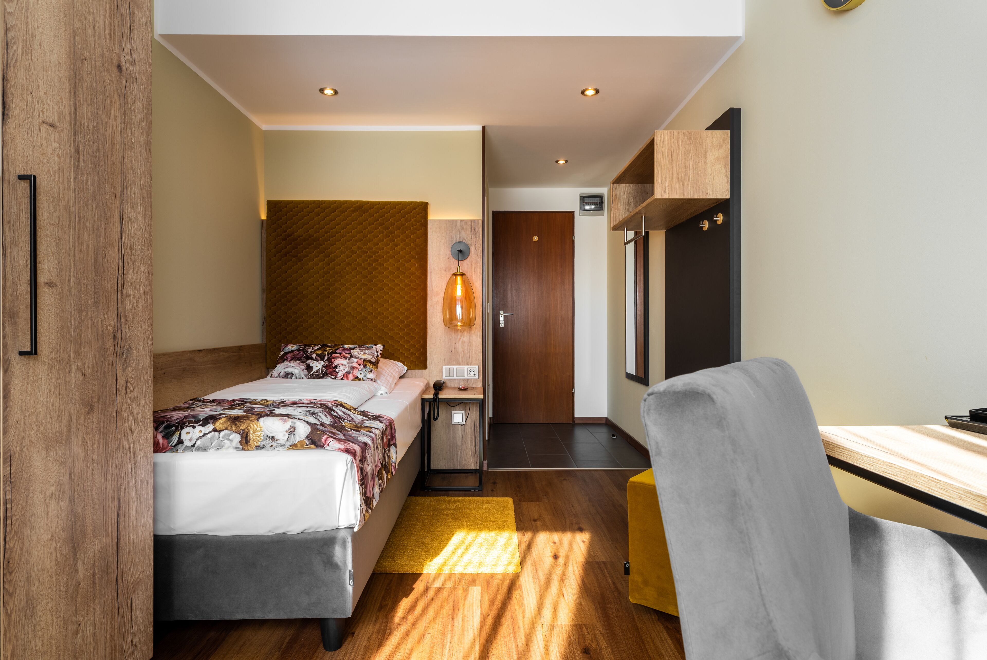 A modern hotel room with a bed, desk, chair, and a small kitchenette.