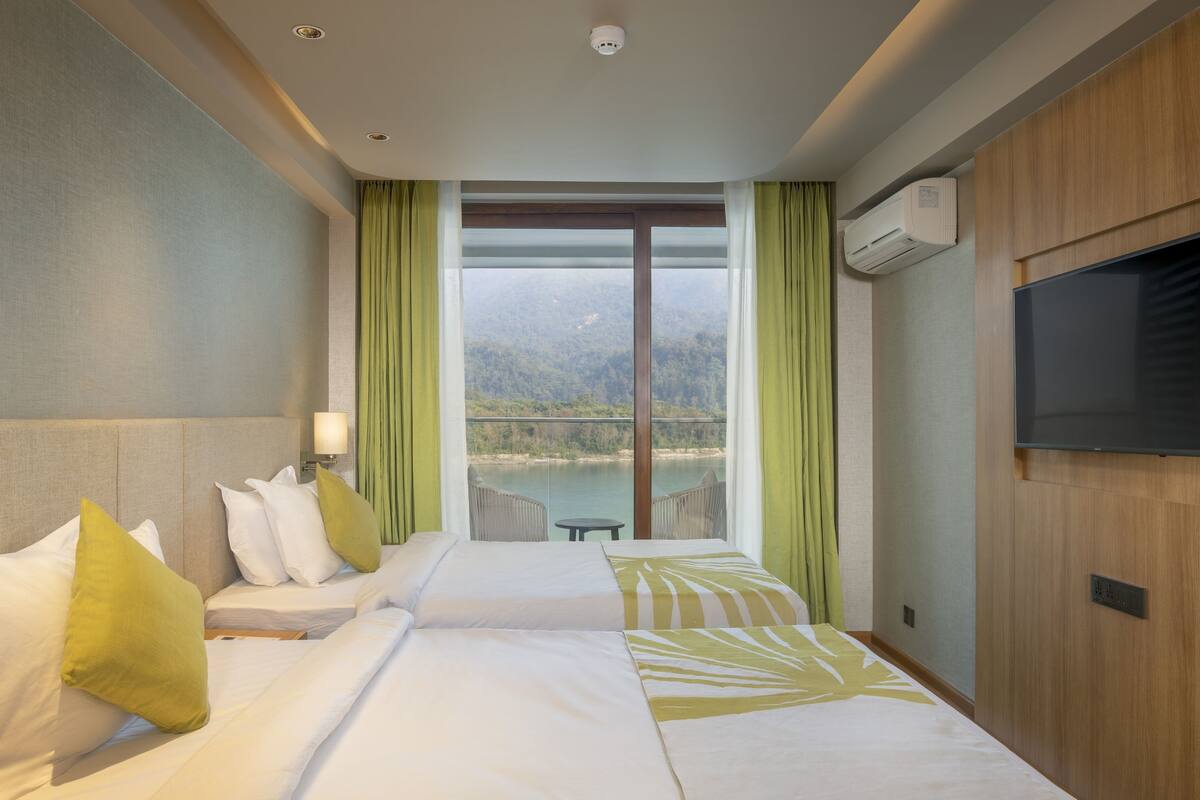 A hotel room with two beds, a TV, an air conditioning unit, and a view of a lake and mountains.