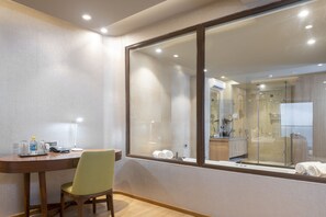 Une chambre d’hôtel avec un bureau en bois, une chaise verte, une grande fenêtre et une douche en verre.