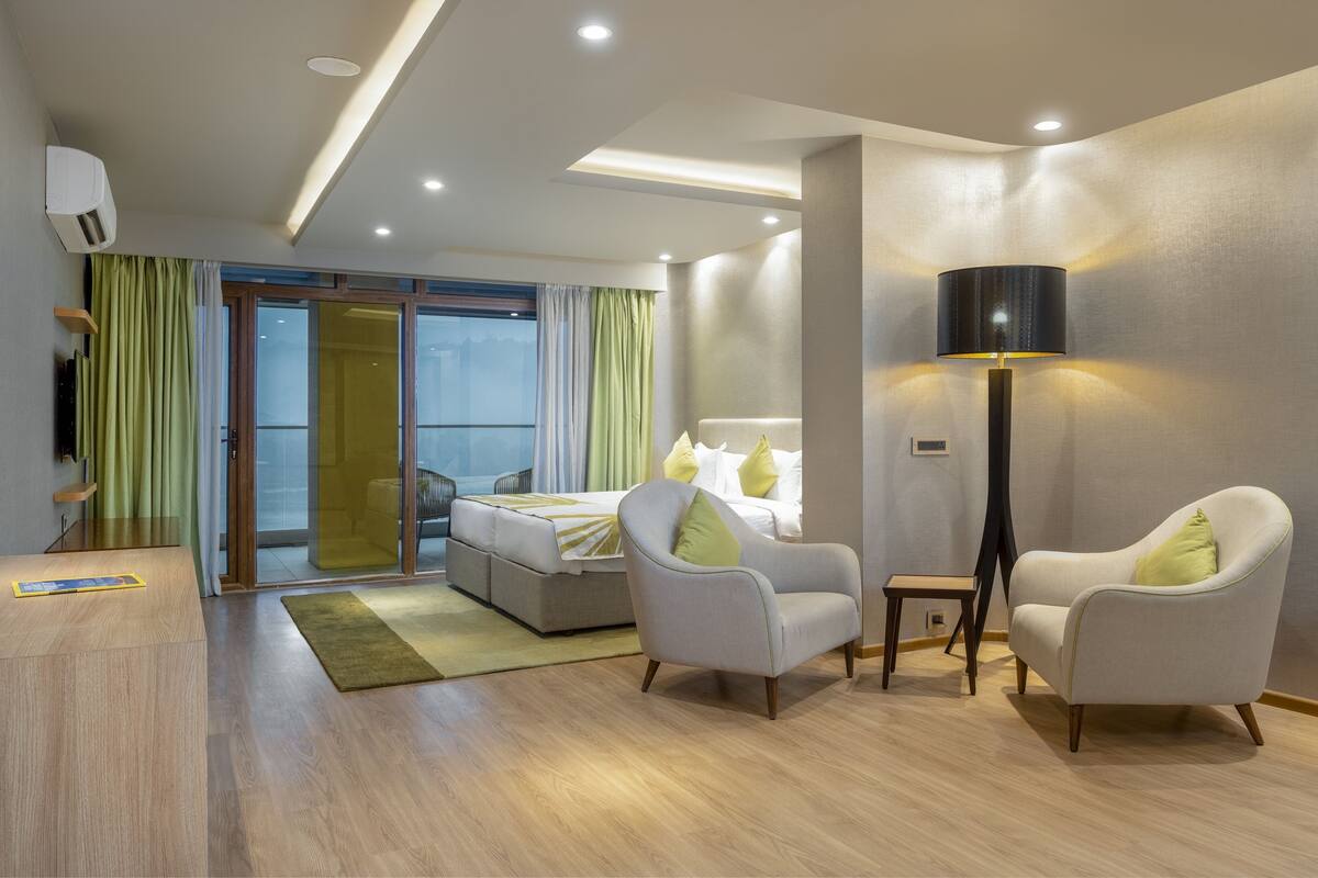 A modern hotel room with a bed, two armchairs, a floor lamp, a desk, and a sliding glass door leading to a balcony.