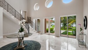 Um hall amplo com uma mesa de centro de vidro, uma escada com guarda-corrimão preto e vista para um jardim através de grandes portas de correr de vidro.