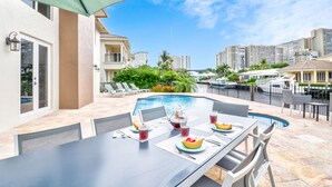 Uma área de refeições à beira da piscina com uma mesa posta para quatro pessoas, uma jarra de chá gelado e uma cesta de frutas.