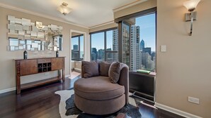 A spacious living room with a large window offering a city view, a round brown sofa, a wooden console table, and a decorative mirror wall. - LOCATION!!  2b/2b Condo, 51st Floor AMAZING Views, Pool and other Amenities! (Chicago)