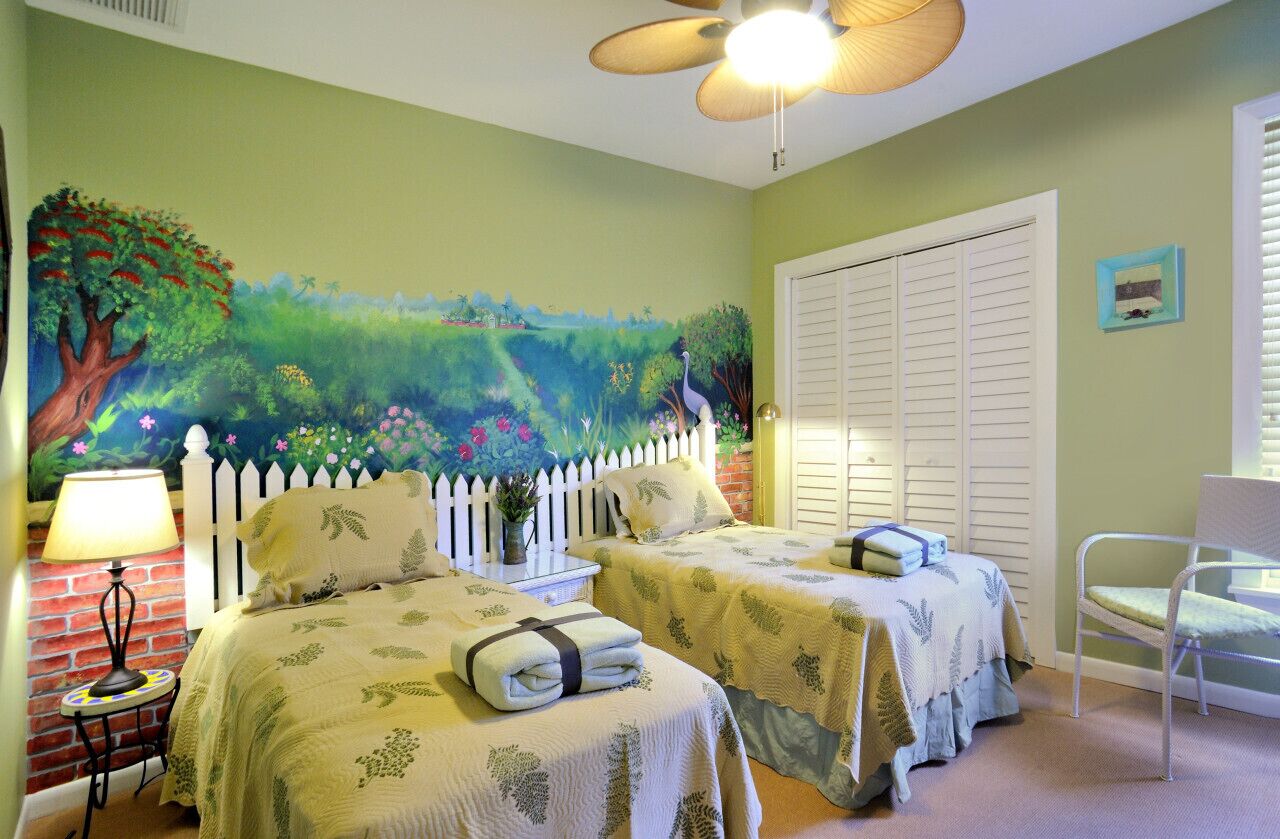 A room with two beds, a white picket fence, a mural of a forest, a ceiling fan, and a chair.